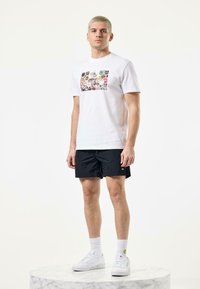 White cotton t-shirt with colorful graphic prints on the front, paired with black swim shorts and white sneakers, standing on a marble pedestal.