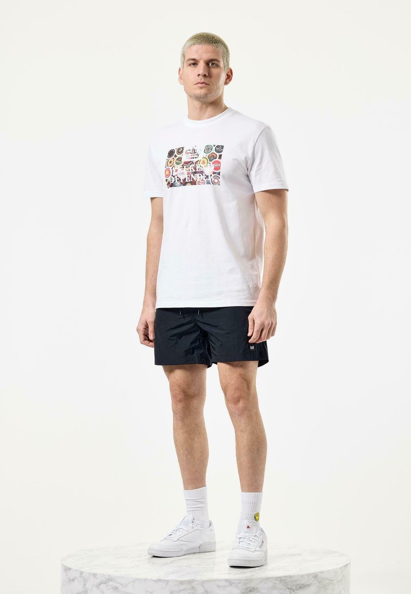 White cotton t-shirt with colorful graphic prints on the front, paired with black swim shorts and white sneakers, standing on a marble pedestal.