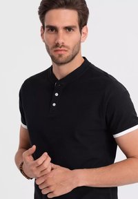 Černé polo tričko s krátkým rukávem z texturované látky, s límečkem a třemi bílými knoflíky, doplněné bílými lemy na rukávech.