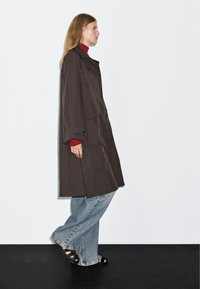 Brązowy oversize'owy płaszcz z dużymi kieszeniami, zapinany na guziki, z szerokimi rękawami, zestawiony z czerwonym golfem, jasnoniebieskimi dżinsami i czarnymi sandałami.