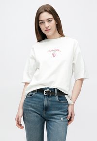 Joven mujer con una camiseta blanca de Calvin Klein y jeans azules con un cinturón negro, de pie frente a un fondo claro y simple.