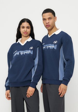 Γυναίκα και άνδρας στέκονται δίπλα-δίπλα φορώντας ταιριαστά μπλε σκούρα αθλητικά πουλόβερ με λευκές γιακάδες και γκρι παντελόνια μπροστά σε μονόχρωμο φόντο.