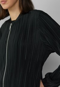 Veste bomber plissée noire avec fermeture éclair à l'avant, tissu texturé et manches longues, présentant une finition lisse et une silhouette structurée.