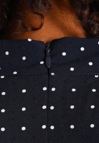 Tessuto nero con pois bianchi, con una zip chiusa sulla parte posteriore del collo e capelli ricci parzialmente visibili sopra.