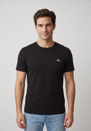 Giovane uomo con capelli corti e scuri che indossa una maglietta nera con un piccolo logo verde sul petto e jeans azzurri, in piedi davanti a uno sfondo semplice.