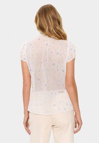 White floral blouse with short puff sleeves, featuring a soft, sheer fabric and colorful flower patterns throughout. Scalloped neckline detail.