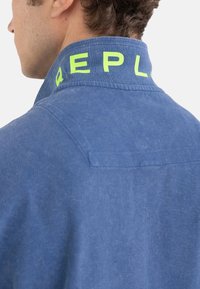 Chaqueta de mezclilla azul con un cuello estructurado que presenta letras de color verde brillante en la banda. Incluye un detalle de bolsillo en la parte posterior.