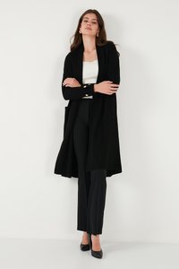 Svart ribbade lång cardigan med öppen front, fickor och knapppdetaljer, bärs över en vit topp och matchas med svarta byxor.