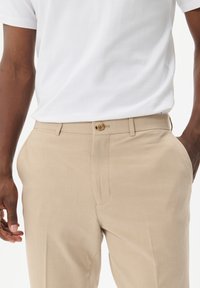 Beige Hose mit glattem Stoffstruktur, ausgestattet mit einer Vordertasche, Gürtelschlaufen und einem braunen Knopfverschluss am Bund.