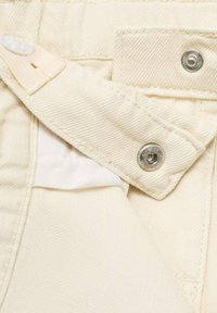 Tissu en denim couleur crème avec une fermeture à bouton et à pression. Présente des coutures visibles et une petite étiquette intérieure. Surface texturée.