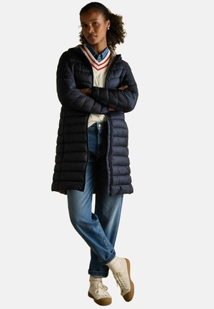 Langer marineblauer Daunenmantel mit gestepptem Design, Reißverschluss vorne und Kapuze. Getragen über einem cremefarbenen Pullover mit roten Streifen, kombiniert mit blauen Jeans und beigen Sneakers.