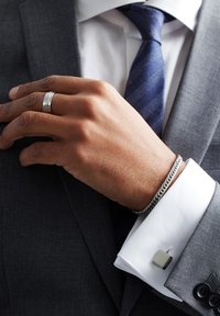 Zilveren armband om pols, grijze blazer, witte shirt met manchetknopen. Hand met een gepolijste zilveren ring, donkerblauwe stropdas met diagonale patroon.