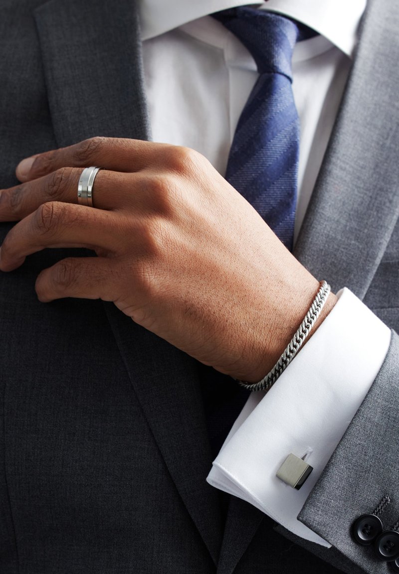 Zilveren armband om pols, grijze blazer, witte shirt met manchetknopen. Hand met een gepolijste zilveren ring, donkerblauwe stropdas met diagonale patroon.