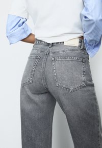 Jeans in denim grigio sbiadito con vita alta, dotati di due tasche posteriori e un'etichetta bianca in vita. Camicia a righe blu e bianche visibile.