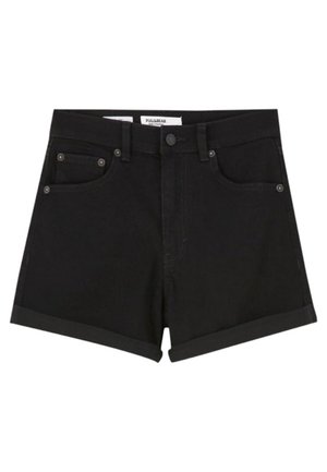 Kvinde iført en sort cropped denimjakke, sorte shorts med opbukket kant og hvide snørebånds-ankelstøvler, stående mod en ensfarvet baggrund.