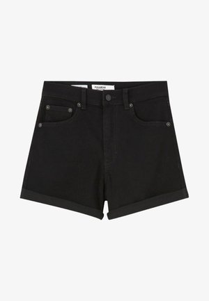 Femme portant une veste en jean noire courte, un short noir à ourlet retroussé et des bottines blanches à lacets, debout devant un fond uni.