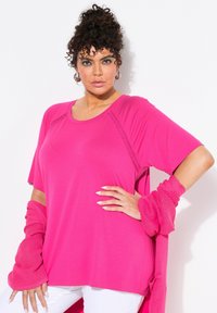 T-shirt braccio corto, morbido e di colore rosa brillante con dettagli testurizzati alle spalle, abbinato a maniche a scrunchy dello stesso colore, indossato sopra pantaloni bianchi.