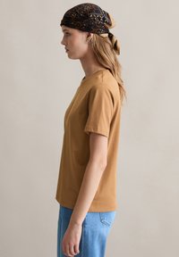 Braunes Kurzarm-T-Shirt mit Rundhalsausschnitt, weicher Textur, getragen mit hellblauen Jeans und einem gemusterten Kopftuch, das hinten gebunden ist.