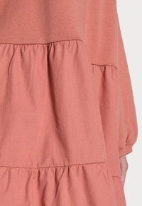 Rypistynyt mekko korallinvärisessä sävyssä, jossa on pehmeä rakenne, rento istuvuus ja pitkät hihat sekä kerätty vyötärö, luoden kerroksellista vaikutelmaa.