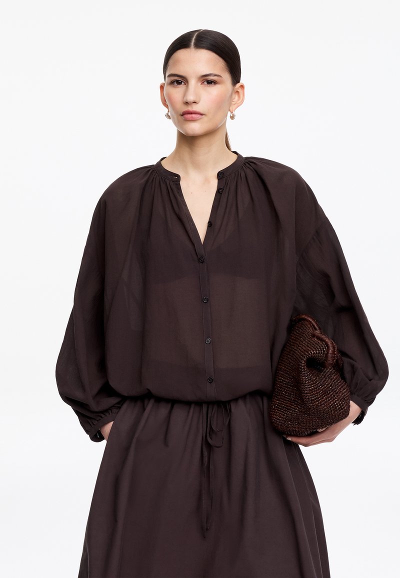 Femme aux cheveux lisses portant un chemisier sombre à boutons et une jupe amples, tenant une pochette tressée marron foncé sur un fond blanc.