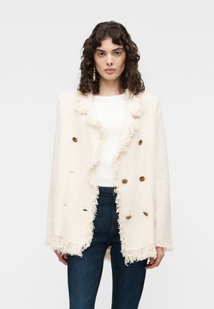 Frau mit lockigem Haar trägt eine cremefarbene, strukturierte zweireihige Jacke mit Fransen-Details über einem weißen Oberteil und dunkelblauen Jeans.