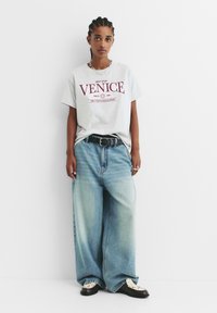 Šedé bavlněné tričko s grafikou "VENICE", kombinované se světle modrými oversized džíny a černými botami s krémovými akcenty. Stříbrný řetízkový náhrdelník.