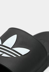 Sandale noire à semelle plate avec une large bride ornée d'un logo à trois feuilles blanc et de trois bandes blanches. Matériau texturé avec une finition lisse.