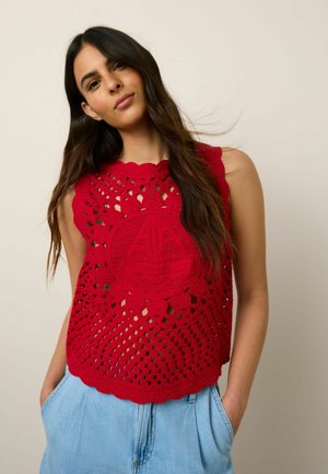 Hosszú sötét hajú nő, piros, horgolt, ujjatlan felsőt visel, amelyen egy vitorlás motívum látható, valamint világoskék, magas derekú nadrágot, kezét a zsebében tartva.