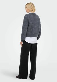 Blonde Frau, die von hinten zu sehen ist, trägt einen strukturierten dunkelgrauen Pullover über einem weißen Shirt, schwarze weite Hosen und dunkle Schuhe vor einem hellen Hintergrund.