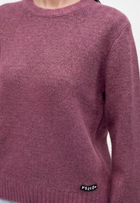 Pull en maille violet à texture douce porté par une personne, avec un petit patch logo Volcom noir et blanc près de l'ourlet.