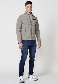 Chaqueta gris claro con cuello alzado, dos bolsillos frontales con cremallera y detalles de costura vertical. Lleva puesta con vaqueros azules y zapatillas blancas.