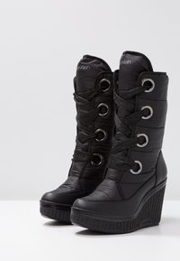 Botas negras de cuña con parte superior acolchada, con ojales plateados, cordones negros y suela de goma texturizada. Logo de la marca en el eje.