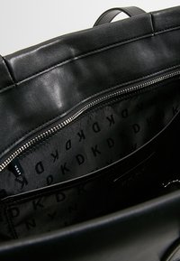 Interiér černé kožené kabelky ukazuje zipovou kapsu s podšívkou z textilie se vzorem, která obsahuje opakující se logo "DKNY" v černé barvě.