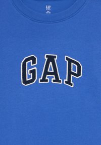 Blå bomullsgenser med rund hals. Har ordet "GAP" i fete, svarte og hvite broderte bokstaver sentrert foran.