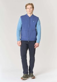 Gilet blu con cerniera e maniche azzurro chiaro, dotato di due tasche frontali. Abbinato a pantaloni scuri e scarpe da ginnastica verdi. Sfondo semplice.