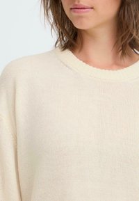 Ljust beige stickad tröja med ribbad rund halsringning och lös passform, med textilstruktur, lätt material och subtila sömnadsdetaljer.