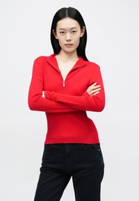 Vörös bordázott, cipzáras pulóver álló gallérral. Testhez simuló szabás és hosszú ujjú. Fekete nadrággal párosítva. Egyszínű háttér.