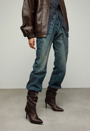 Veste bombardier en cuir marron, jean bleu à finition délavée et bottes marron souples à talons hauts avec des bouts pointus.