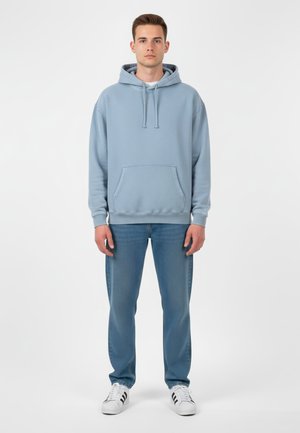 Felpa azzurra in tessuto morbido, con tasca frontale e cappuccio con cordino, abbinata a jeans azzurri e sneakers bianche.
