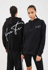 Zwei Personen mit geflochtenem Haar tragen schwarze Kapuzenpullover mit weißem "Your Stern"-Text, einer steht frontal und einer zeigt die Rückansicht.