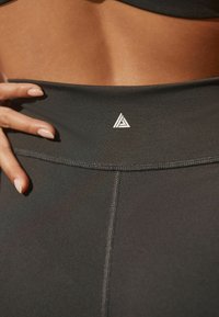Mustat korkeavyötäröiset legginsit, joissa on sileä pinta, valkoisella kolmio-logolla ja kontrastiväriset ompeleet saumoissa.