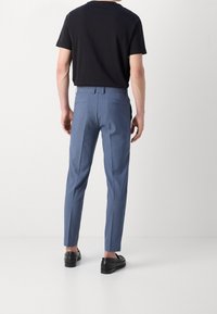 Modré kalhoty na míru slim fit, s dvěma zadními kapsami a hladkým povrchem, v kombinaci s černým tričkem s krátkým rukávem a botami.