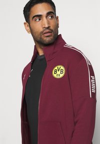 Bordowa kurtka na zamek błyskawiczny z kieszeniami bocznymi, z okrągłym żółtym logo (\