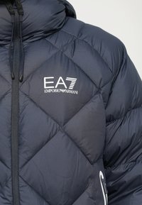 Közelkép egy sötétkék steppelt kabátról, amelyen az EA7 Emporio Armani logó látható, valamint fekete cipzárral és zsinórral ellátott gallérral.