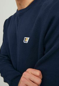 Dunkelblau gestrickter Pullover mit Rundhalsausschnitt, gerippten Bündchen und einem kleinen, bunten Logo-Patch mit einer Jahreszahl, strukturiertem Stoff.