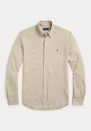 Man wearing beige button-up shirt, white pants, brown belt, watch, and brown loafers, standing with one hand in pocket.