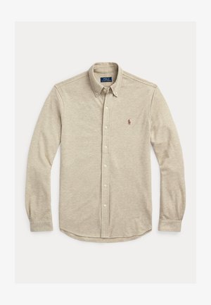 Chemise beige à manches longues boutonnée avec col boutonné et petit logo brodé sur la poitrine gauche, posée à plat sur fond blanc.