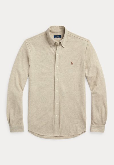 Bež srajca z dolgimi rokavi na gumbe z ovratnikom na gumbe in majhno vezeno značko na levi strani prsnega dela, položena na ravno belo ozadje.