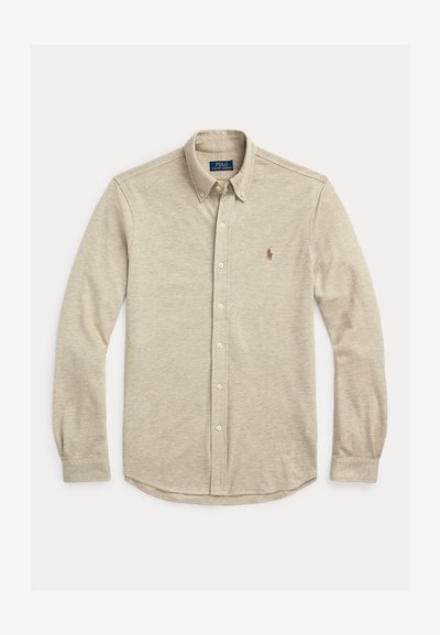 Bež srajca z dolgimi rokavi na gumbe z ovratnikom na gumbe in majhno vezeno značko na levi strani prsnega dela, položena na ravno belo ozadje.