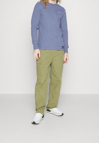 Personne portant une chemise à manches longues bleue, un pantalon cargo vert olive et des baskets blanches, se tenant contre un fond clair uni.